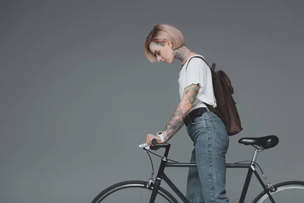 Vue latérale de jeune femme avec sac à dos debout avec vélo isolé sur gris — Photo de stock