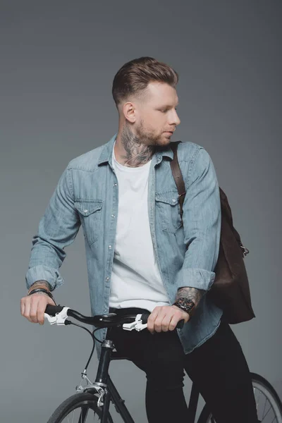 Élégant jeune homme avec sac à bandoulière assis sur le vélo et regardant loin isolé sur gris — Photo de stock
