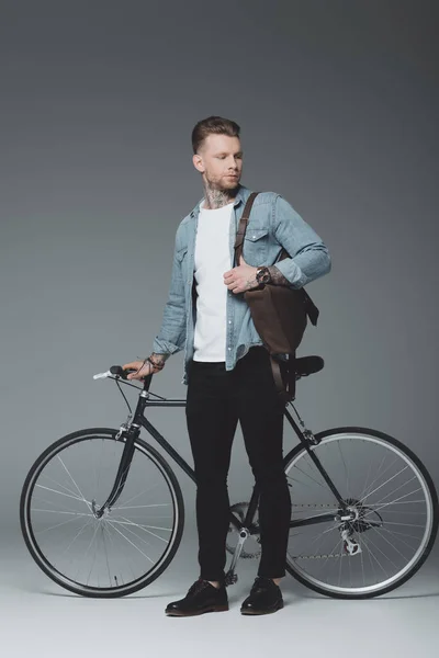 Vue pleine longueur du jeune homme tatoué debout avec vélo et regardant loin sur gris — Photo de stock