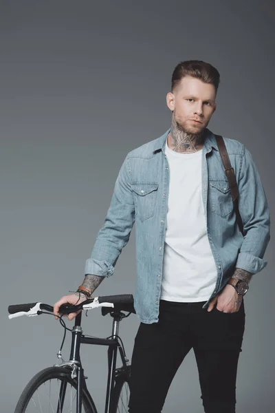 Bel giovanotto con borsa a tracolla e bicicletta in piedi e guardando la fotocamera isolata sul grigio — Foto stock