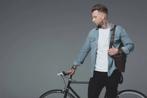 Handsome young man with shoulder bag and bicycle standing isolated on grey — Stock Photo