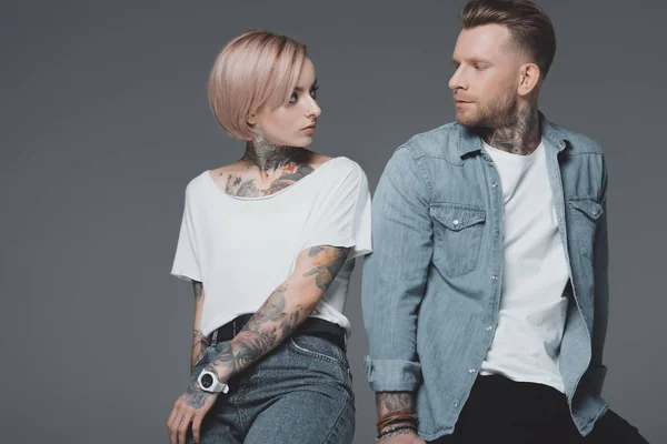 Стильна молода пара з татуюваннями дивиться один на одного ізольовано на сірому — стокове фото