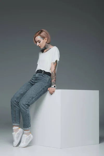 Hermosa joven con tatuajes sentado en cubo y mirando hacia abajo en gris - foto de stock