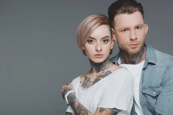 Hermosa joven pareja tatuada en el amor de pie juntos y mirando a la cámara aislada en gris - foto de stock