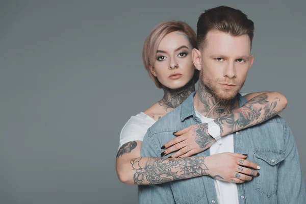 Elegante pareja joven con tatuajes abrazando y mirando a la cámara aislada en gris - foto de stock