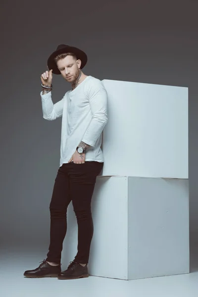 Visão comprimento total do jovem elegante em chapéu com tatuagens em pé perto de cubos brancos em cinza — Fotografia de Stock