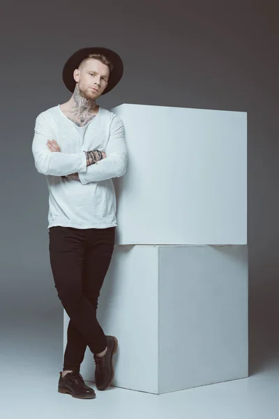 Stylish young man with tattoos standing with crossed arms and looking at camera on grey — Stock Photo