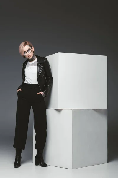 Fashionable young woman in leather jacket and eyeglasses standing with hands in pockets and looking at camera in studio — Stock Photo