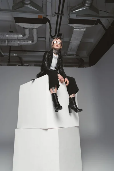 Low angle view of stylish young tattooed woman sitting on white cubes in studio — Stock Photo