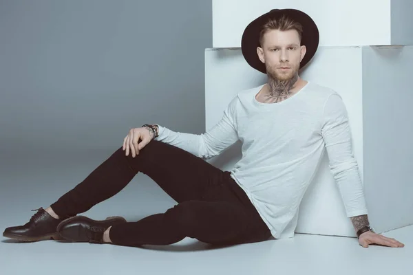 Stylish tattooed man in hat sitting near white cubes, isolated on grey — Stock Photo