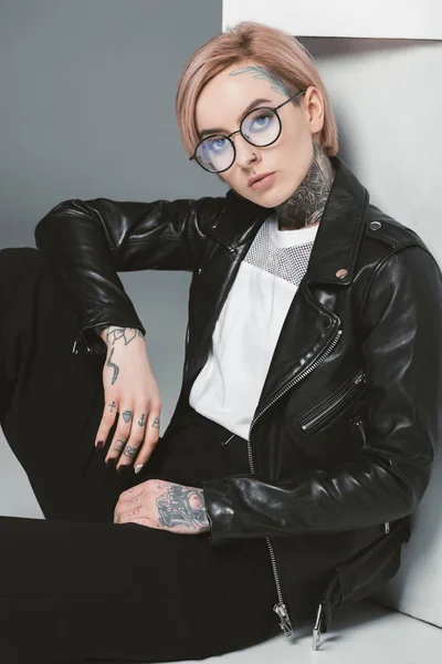 Стильная татуированная девушка в очках и кожаной куртке, сидящая рядом с белыми кубиками, изолированная на сером — стоковое фото
