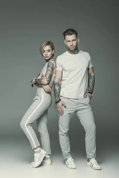 Модна татуйована пара в спортивному одязі позує разом, ізольована на сірому — стокове фото