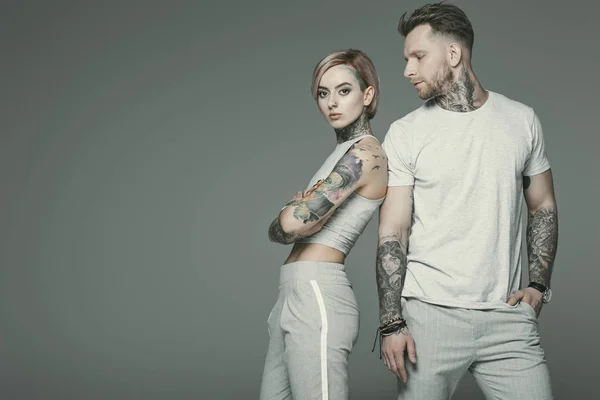 Elegante pareja tatuada en ropa deportiva posando juntos, aislado en gris - foto de stock