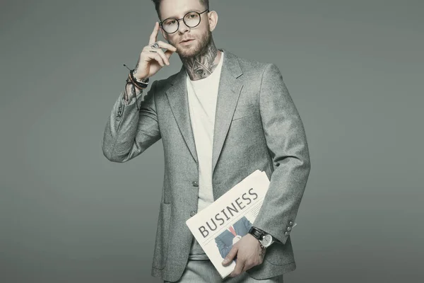 Handsome businessman in eyeglasses and suit holding business newspaper, isolated on grey — Stock Photo