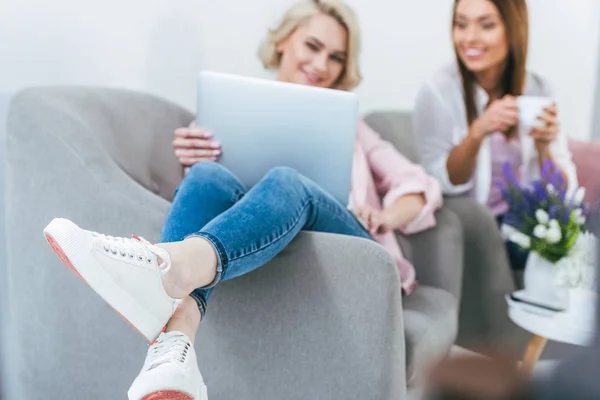 Selective focus of girl using laptop while her friend holding cup of coffee — Stock Photo