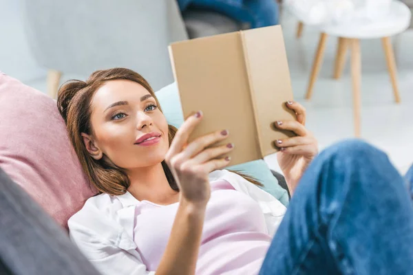 Atraente menina leitura livro enquanto deitado no sofá em casa — Fotografia de Stock