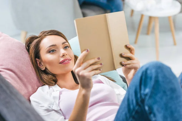 Attrayant livre de lecture fille tout allongé sur le canapé à la maison — Photo de stock