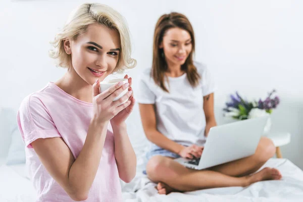 Caucasian girls with coffee and laptop on bed — Stock Photo