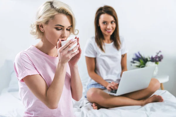 Подруги пьют кофе и пользуются ноутбуком на кровати — стоковое фото