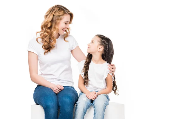 Mother and daughter looking at each other while sitting on white cube isolated on white — Stock Photo