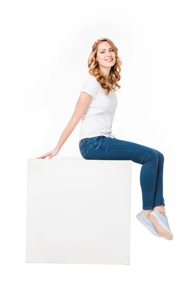 Cheerful woman sitting on empty  white cube isolated on white — Stock Photo