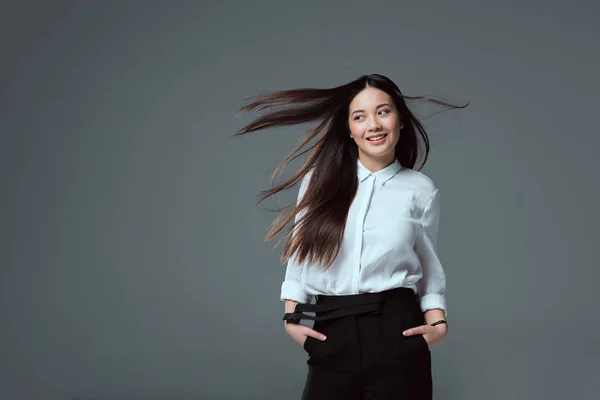 Красива усміхнена азіатська дівчина стоїть з руками в кишенях і дивиться ізольовано на сірому — стокове фото