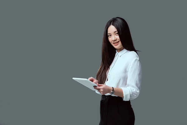 Усміхнена молода азіатська жінка використовує цифровий планшет і дивиться на камеру ізольовано на сірому — стокове фото
