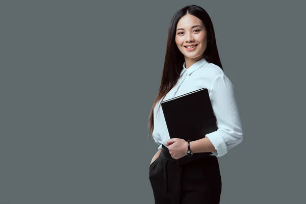 Beautiful young asian businesswoman holding folder and smiling at camera isolated on grey — Stock Photo