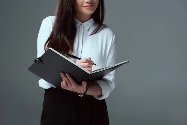 Обрізаний знімок усміхненої молодої бізнес-леді робить нотатки ізольовані на сірому — стокове фото