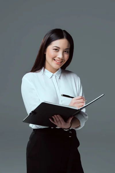 Sorridente giovane donna d'affari prendere appunti e guardando la fotocamera isolata su grigio — Foto stock