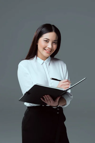 Sonriente joven empresaria tomando notas y mirando a la cámara aislada en gris - foto de stock