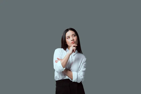 Задумчивая молодая женщина стоит с рукой на подбородке и смотрит в сторону изолированы на серый — стоковое фото