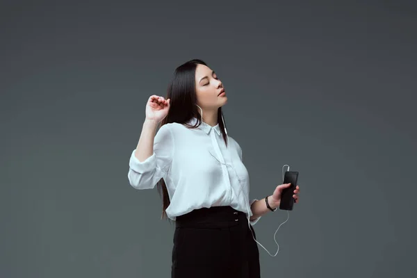 Hermosa tierna joven en auriculares escuchando música con teléfono inteligente aislado en gris - foto de stock