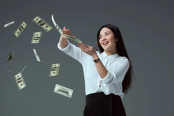 Smiling asian girl throwing dollar banknotes isolated on grey — Stock Photo