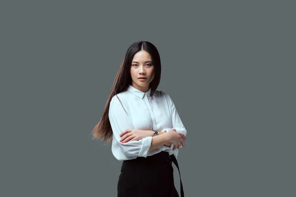 Красива азіатська дівчина стоїть з схрещеними руками і дивиться на камеру ізольовано на сірому — стокове фото