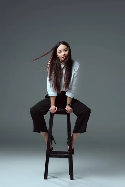 Beautiful young asian woman sitting on chair and looking at camera on grey — Stock Photo