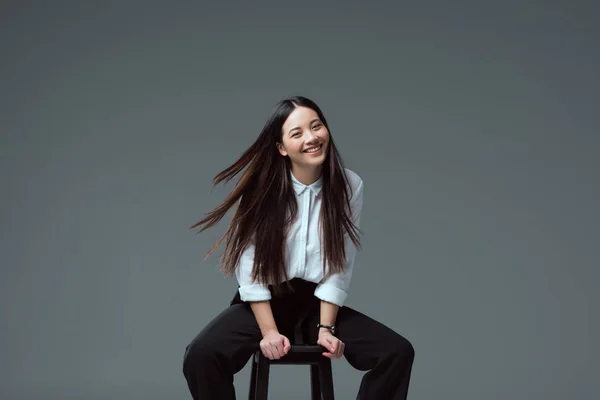 Привлекательная азиатская девушка, сидящая на стуле и улыбающаяся в камеру, изолированную на сером — стоковое фото
