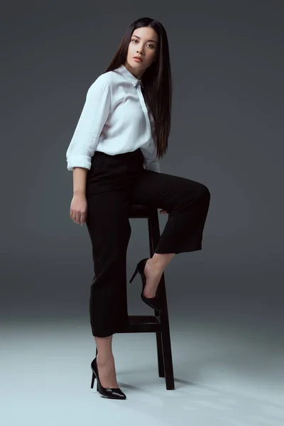 Full length view of beautiful stylish asian girl posing with chair and looking at câmera on grey — Fotografia de Stock