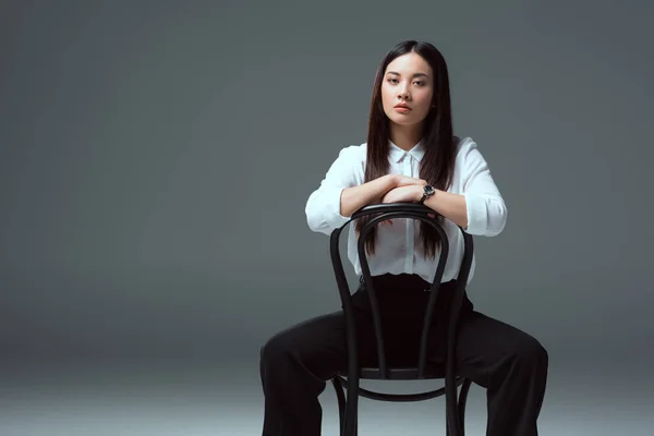 Schön asiatische Mädchen sitzen auf Stuhl und Blick auf Kamera auf grau — Stockfoto