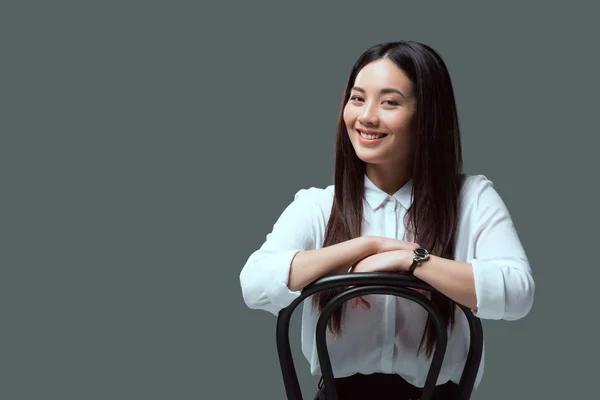 Привлекательная молодая азиатская женщина, сидящая на стуле и улыбающаяся в камеру, изолированную на сером — стоковое фото