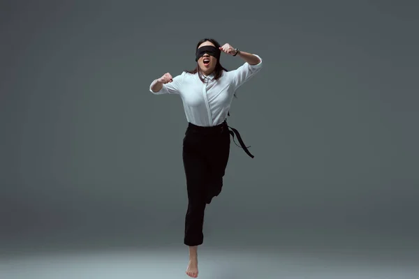 Aggressive young barefoot woman in black blindfold jumping with open mouth on grey — Stock Photo