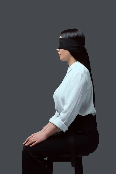 Side view of young blindfolded woman sitting on chair isolated on grey — Stock Photo