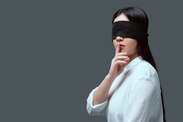 Mujer joven con los ojos vendados negro gesto de silencio aislado en gris - foto de stock