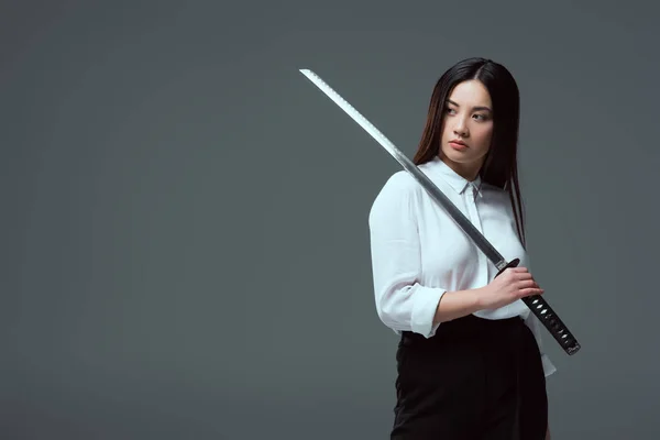 Приваблива молода азіатська жінка тримає меч катани і дивиться ізольовано на сірому — стокове фото
