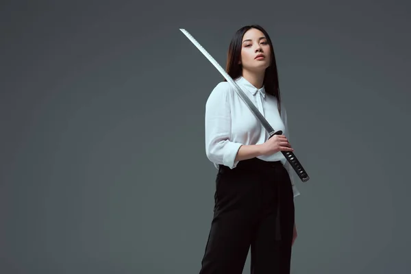 Приваблива молода азіатська жінка тримає меч катани і дивиться на камеру ізольовано на сірому — стокове фото