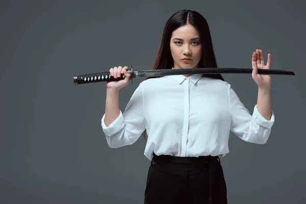 Beautiful asian girl holding katana sword and looking at camera isolated on grey — Stock Photo