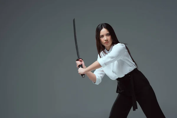 Joven asiático mujer luchando con katana espada y mirando a cámara aislado en gris - foto de stock