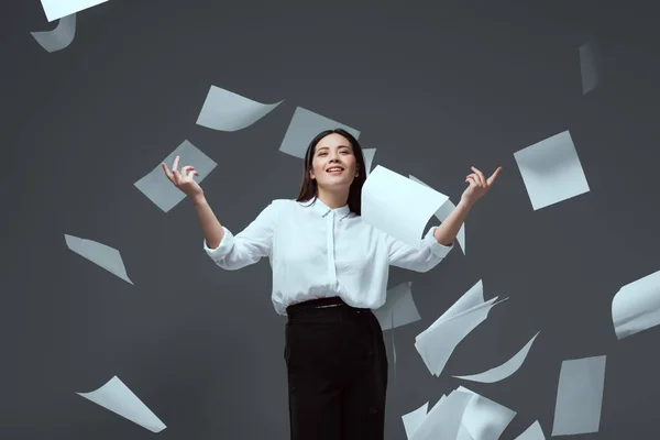 Lächelnde junge Geschäftsfrau wirft Papiere isoliert auf grau — Stockfoto