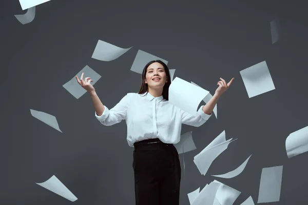 Sonriente joven empresaria lanzando papeles aislados en gris - foto de stock