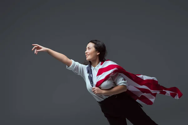 Glückliche junge Frau mit amerikanischer Flagge, die Hand ausstreckt und isoliert auf grau wegschaut — Stockfoto